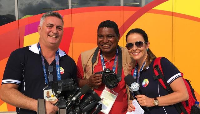 Após entrevista para o jornal do SBT, à direita a repórter cor de pele parda, cabelos lisos amarrados, trajando uma camisa polo preta e calça rosa pink com uma mochila vermelha em suas costas, usando também óculos escuro, carregando em suas mão o microfone da emissora. A esquerda o repórter cinematográfico, cor de pele branca, cabelos grisalhos trajando uma camisa polo preta, calça jeans preta carregando em suas mão uma câmera de vídeo, todos com expressão de felicidade.